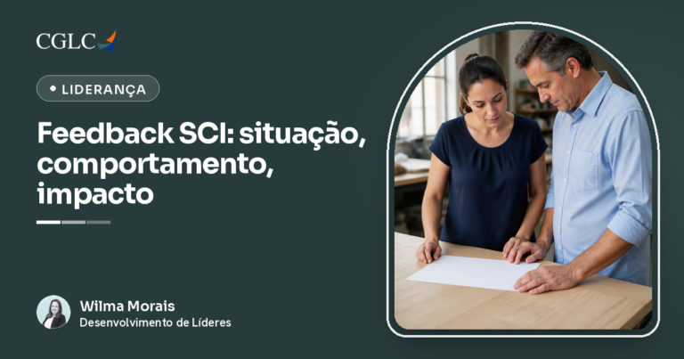 Mulher de cabelo preso e blusa azul-marinho ao lado de um homem grisalho de camisa azul-clara com as mangas dobradas, os dois em pé, inclinados sobre uma mesa de madeira clara, olhando juntos para uma folha de papel em branco, com as mãos apoiadas na mesa.
