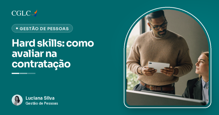 Homem de óculos e suéter bege, em pé junto à janela, examinando um tablet que segura com as duas mãos; sentada ao lado, uma mulher loura de blazer escuro olha para ele; ao fundo, uma sala de escritório e um monitor de costas