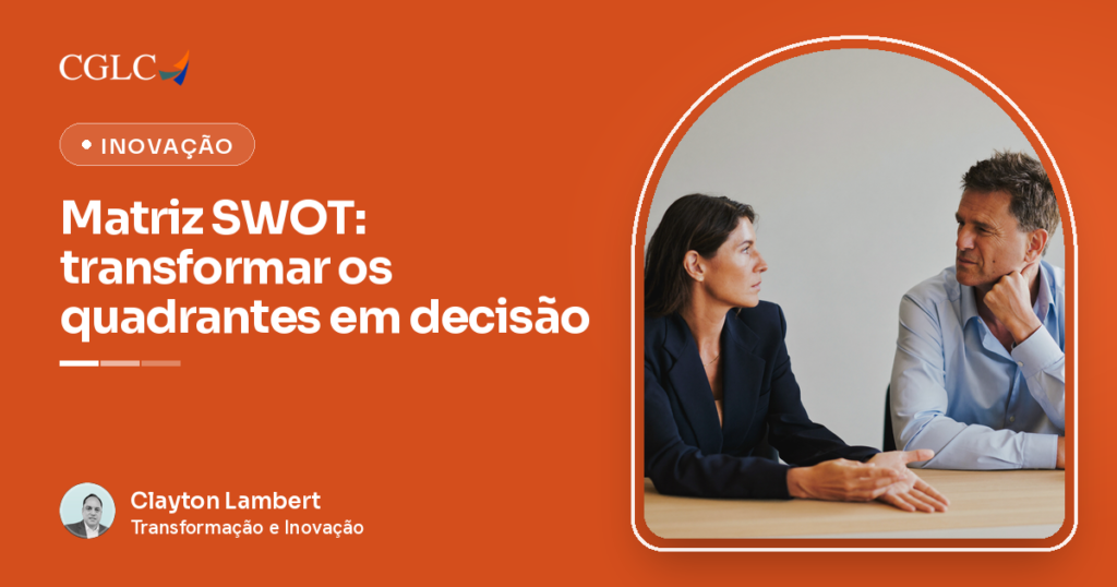 Mulher de blazer escuro e homem de camisa azul-clara sentados um de frente para o outro a uma mesa de madeira clara, ela com a palma da mão aberta apoiada no tampo enquanto fala e ele ouvindo com a mão no queixo.